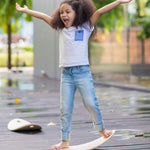A child standing on the PlanToys Balance Board with both their arms help up high in a y shape. The board has been positioned on wooden decking outdoors. 