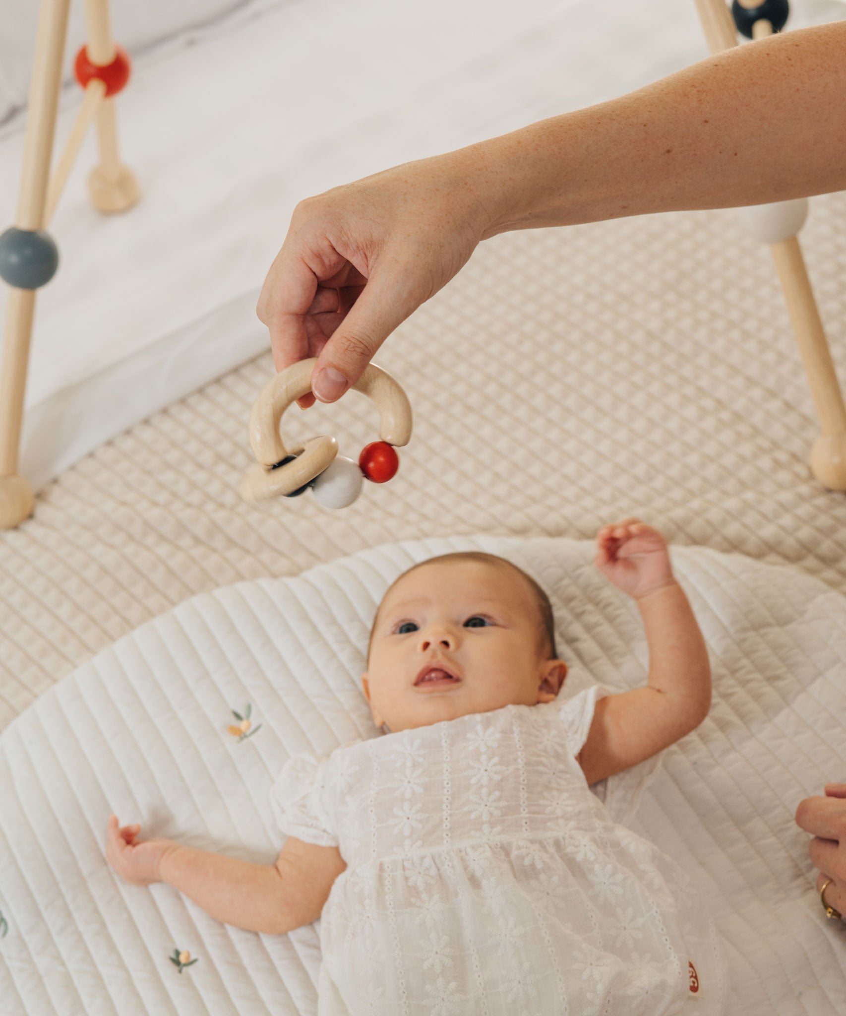 A close up of an adult's hand holding up a PlanToys Beads Rattle in a High Contrast colourway above a baby. 
PlanToys range of toys are made from sustainably sourced rubberwood, we're proud stockists of PlanToys here at Babipur. 