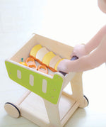 A PlanToys Shopping Trolley on a white surface, a child's hand can be seen pushing the trolley along. 