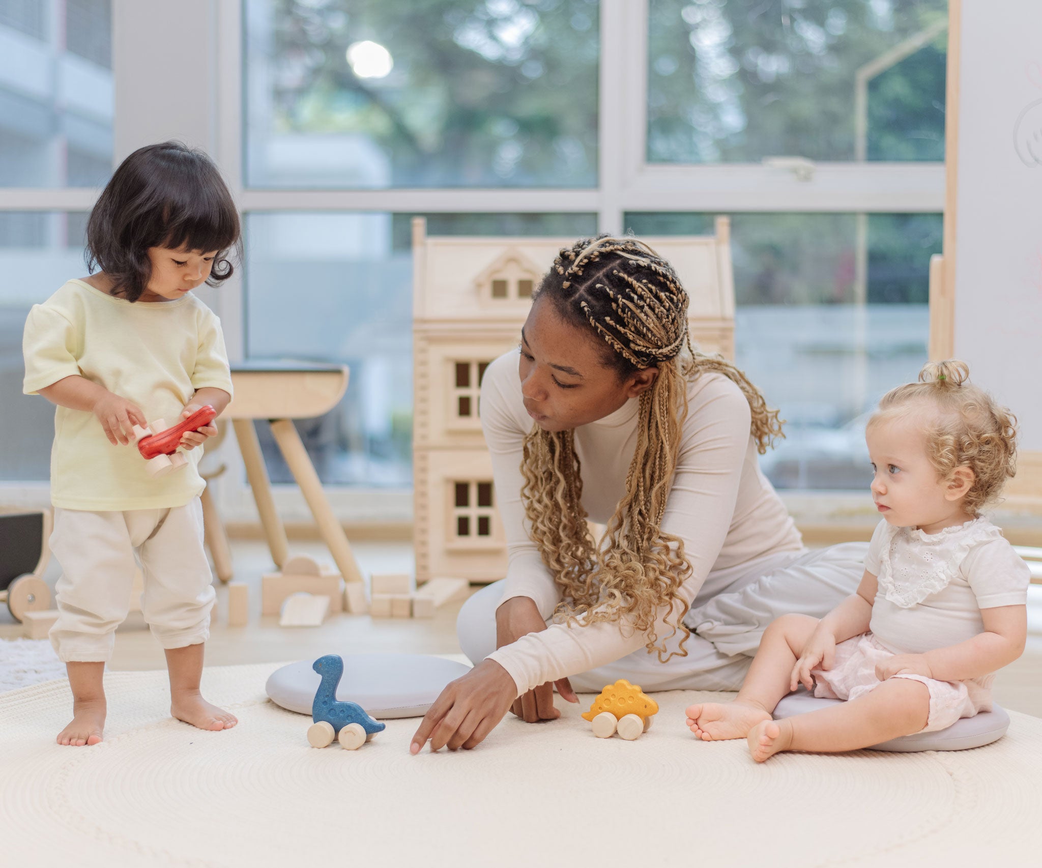 Two children and an adult playing with the PlanToys Dino wooden Car toys. A blue coloured diplodocus, a yellow stegosaurus and red coloured t-rex dinosaur figures on wheels. PlanToys range of toys are made from sustainably sourced rubberwood, we're proud stockists of PlanToys here at Babipur. 