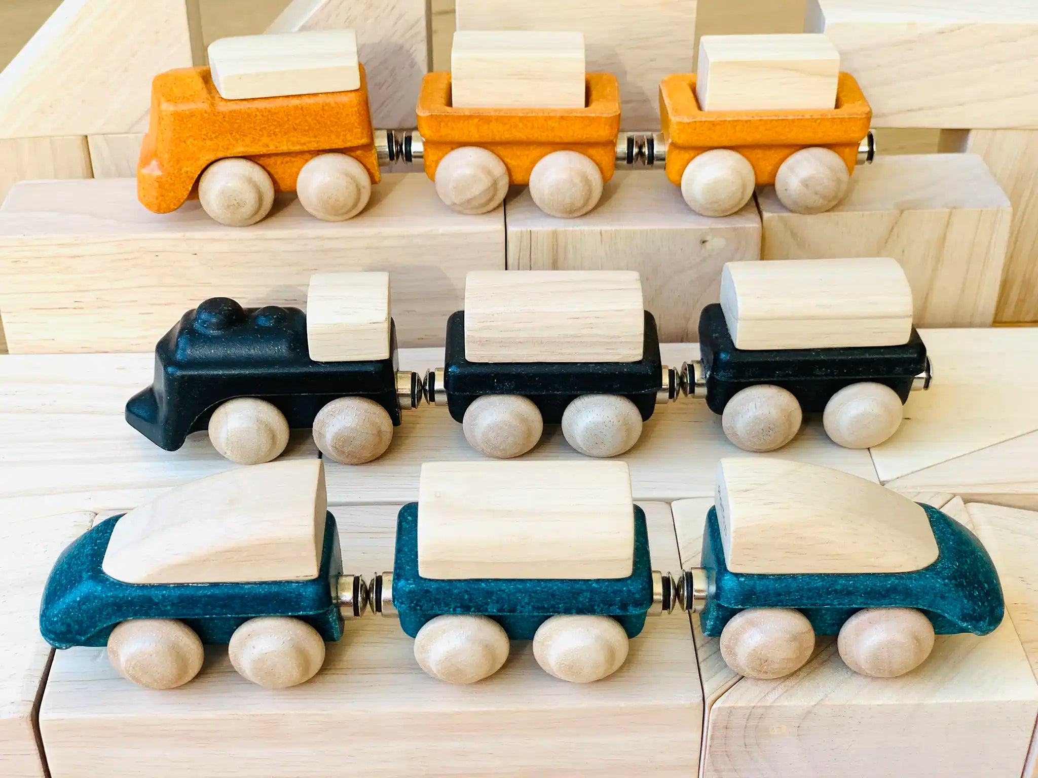Three PlanToys wooden trains placed on wooden blocks. One yellow, one black and one blue. 