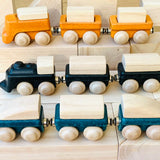 Three PlanToys wooden trains placed on wooden blocks. One yellow, one black and one blue. 