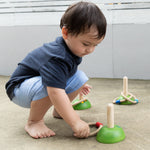 A child playing with the PlanToys Meadow Ring Toss they are kneeling down picking up one of the rings off the ground. 
