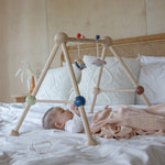 A baby laying underneath the PlanToys Play Gym Orchard on a bed which is covered in crisp white linen. 