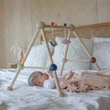 A baby laying underneath the PlanToys Play Gym Orchard on a bed which is covered in crisp white linen. 