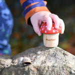 A child's hand reaching for a red PlanToys Mushroom Kaleidoscope that has been placed on a large rock. 