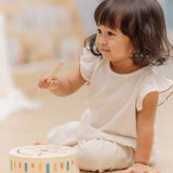 Child playing with the PlanToys wooden sensory drum musical toy with the wood drumstick in hand.