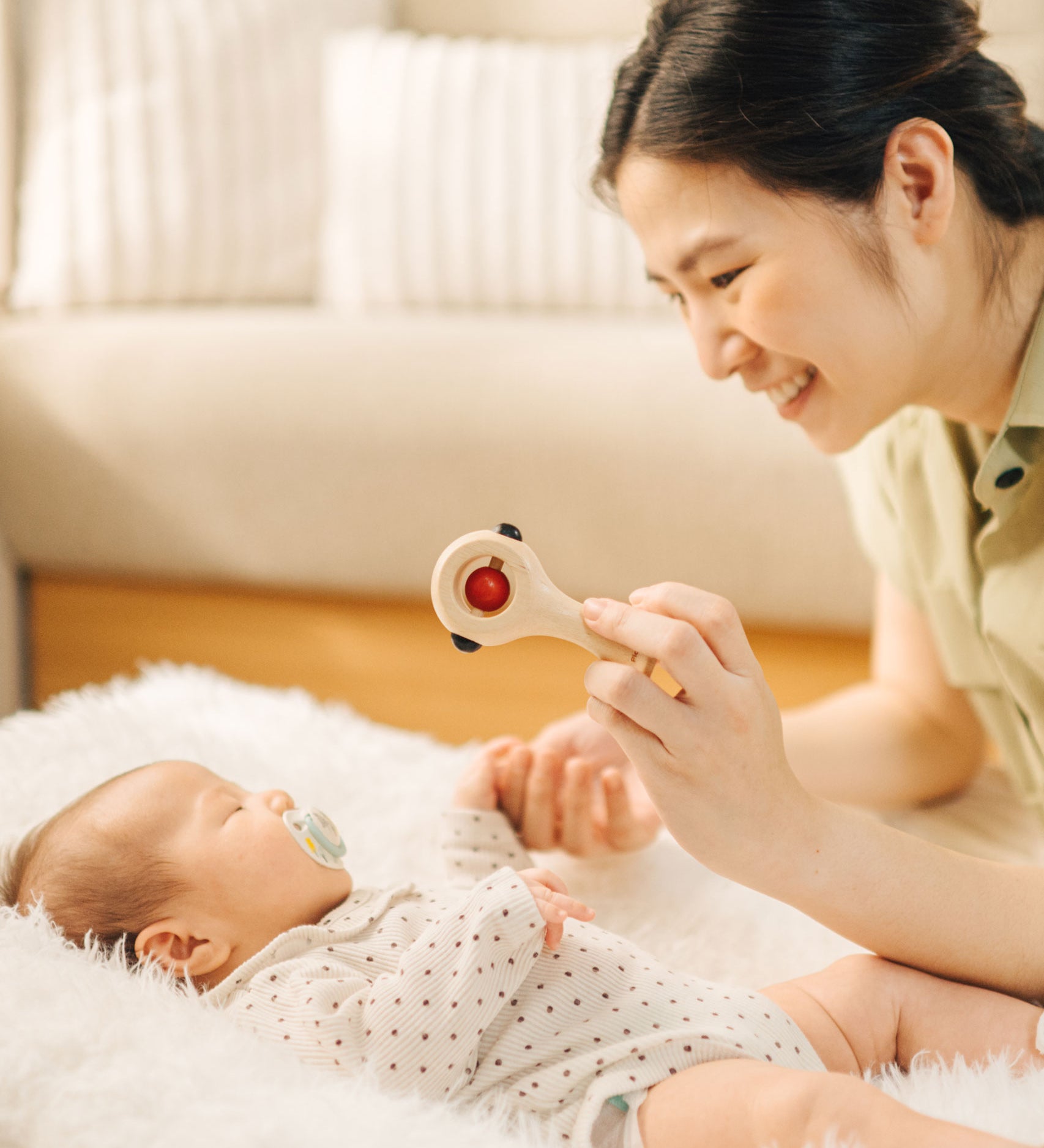 An adult holding up the PlanToys Shaking Rattle in a High Contrast colourway above a baby. The red and black details stand out against the natutral rubberwood body. PlanToys range of toys are made from sustainably sourced rubberwood, we're proud stockists of PlanToys here at Babipur. 