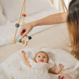 An adult holding the PlanToys high contrast square clutching toy above a baby that is laying underneath a baby gym. PlanToys range of toys are made from sustainably sourced rubberwood, we're proud stockists of PlanToys here at Babipur. 