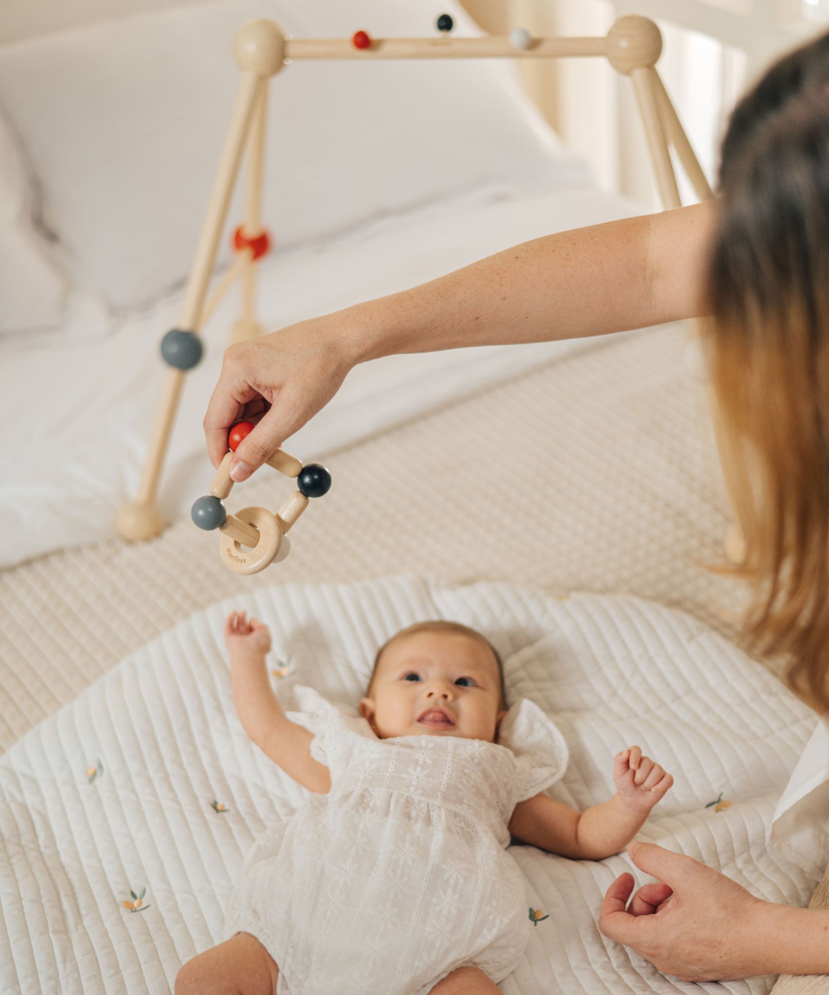 An adult holding the PlanToys high contrast square clutching toy above a baby that is laying underneath a baby gym. PlanToys range of toys are made from sustainably sourced rubberwood, we're proud stockists of PlanToys here at Babipur. 