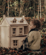 A child sitting on a leaf covered floor in the forest playing with a PlanToys Victorian Dolls House.
