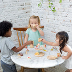Two children at a table having a pretend tea party with the PlanToys bakery Stand in the centre.