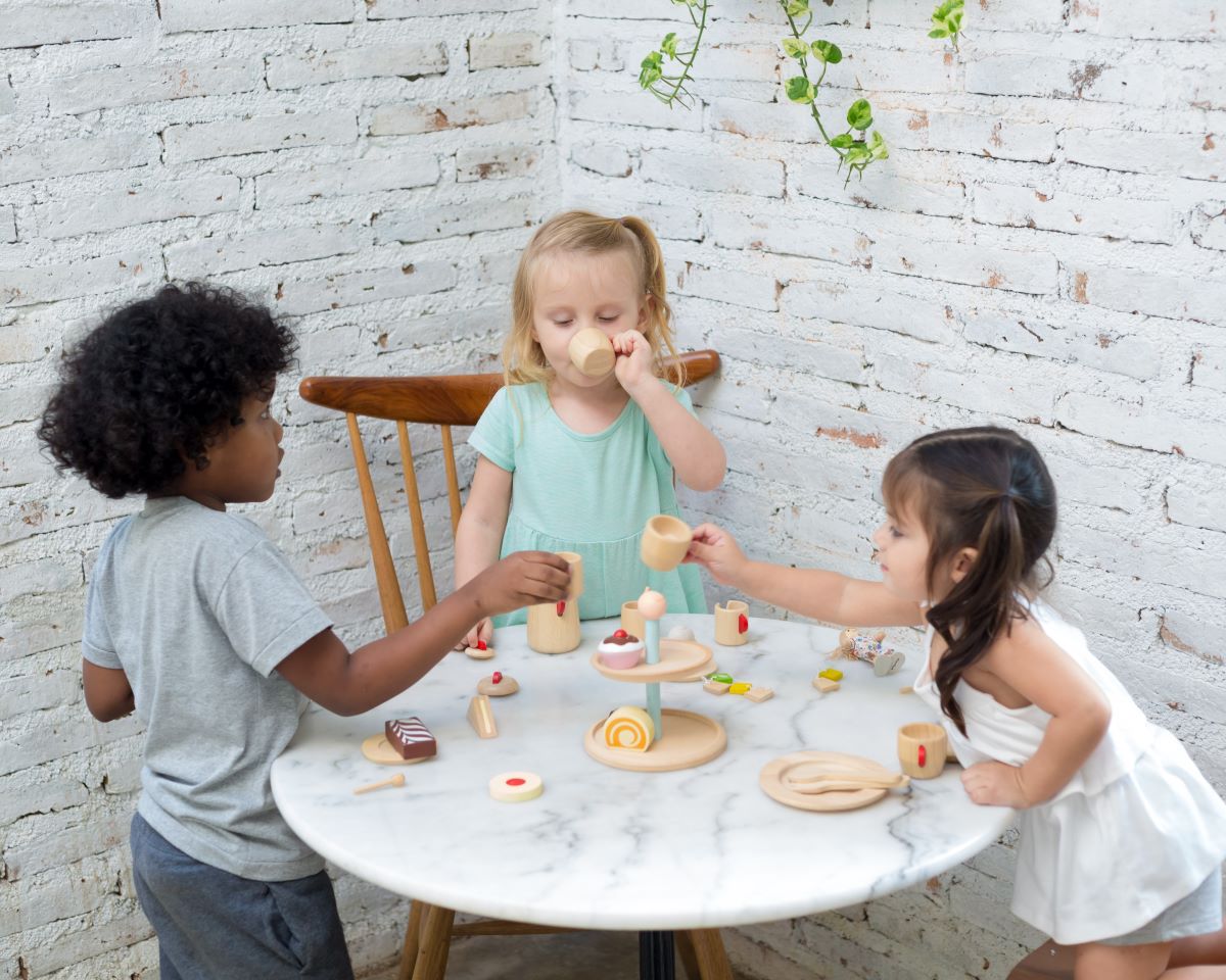 Two children at a table having a pretend tea party with the PlanToys bakery Stand in the centre.