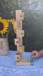 A child playing with the Beck Toys wooden roller cup marble run, outside in the Babipur garden. As he places marbles on the top, they drop into little wooden cups, which tip and drop the marble down into the next cup. 