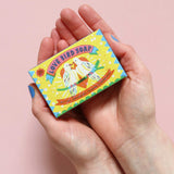 Printed Peanut love bird natural handmade soap with tea tree lemongrass lavender & peppermint. The bar is in decorative packaging in a person's hands.
