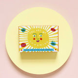The Printed Peanut sunshine natural handmade orange and grapefruit soap. The bar is shown in a decorative packaging box placed on a yellow circular surface. 