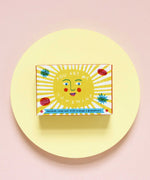 The Printed Peanut sunshine natural handmade orange and grapefruit soap. The bar is shown in a decorative packaging box placed on a yellow circular surface. 