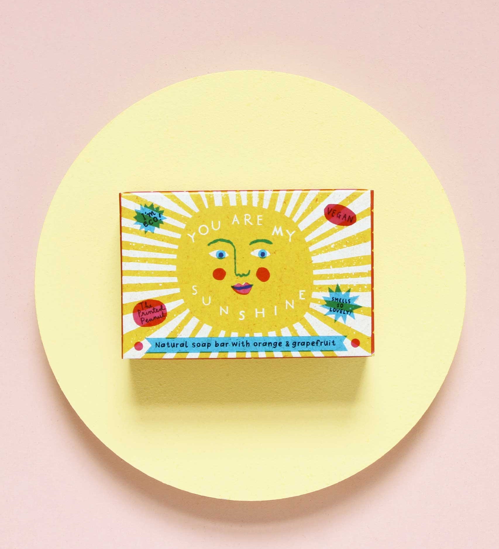The Printed Peanut sunshine natural handmade orange and grapefruit soap. The bar is shown in a decorative packaging box placed on a yellow circular surface. 