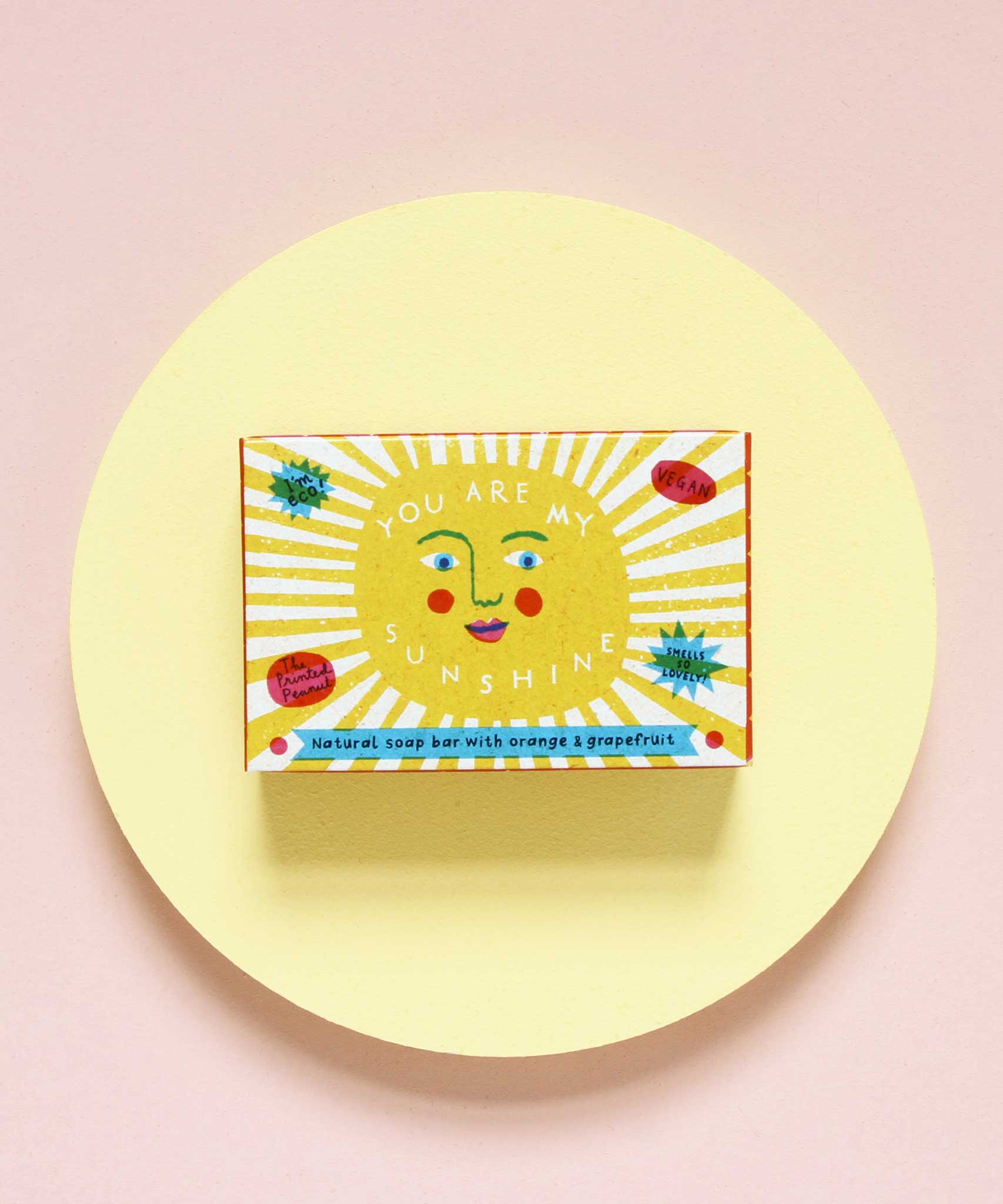 The Printed Peanut sunshine natural handmade orange and grapefruit soap. The bar is shown in a decorative packaging box placed on a yellow circular surface. 