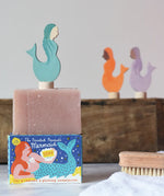 Printed Peanut rose geranium natural soap bar placed on top of the mermaid themed illustrated packaging box. Grimm's mermaid decorative figure has been placed on top. 