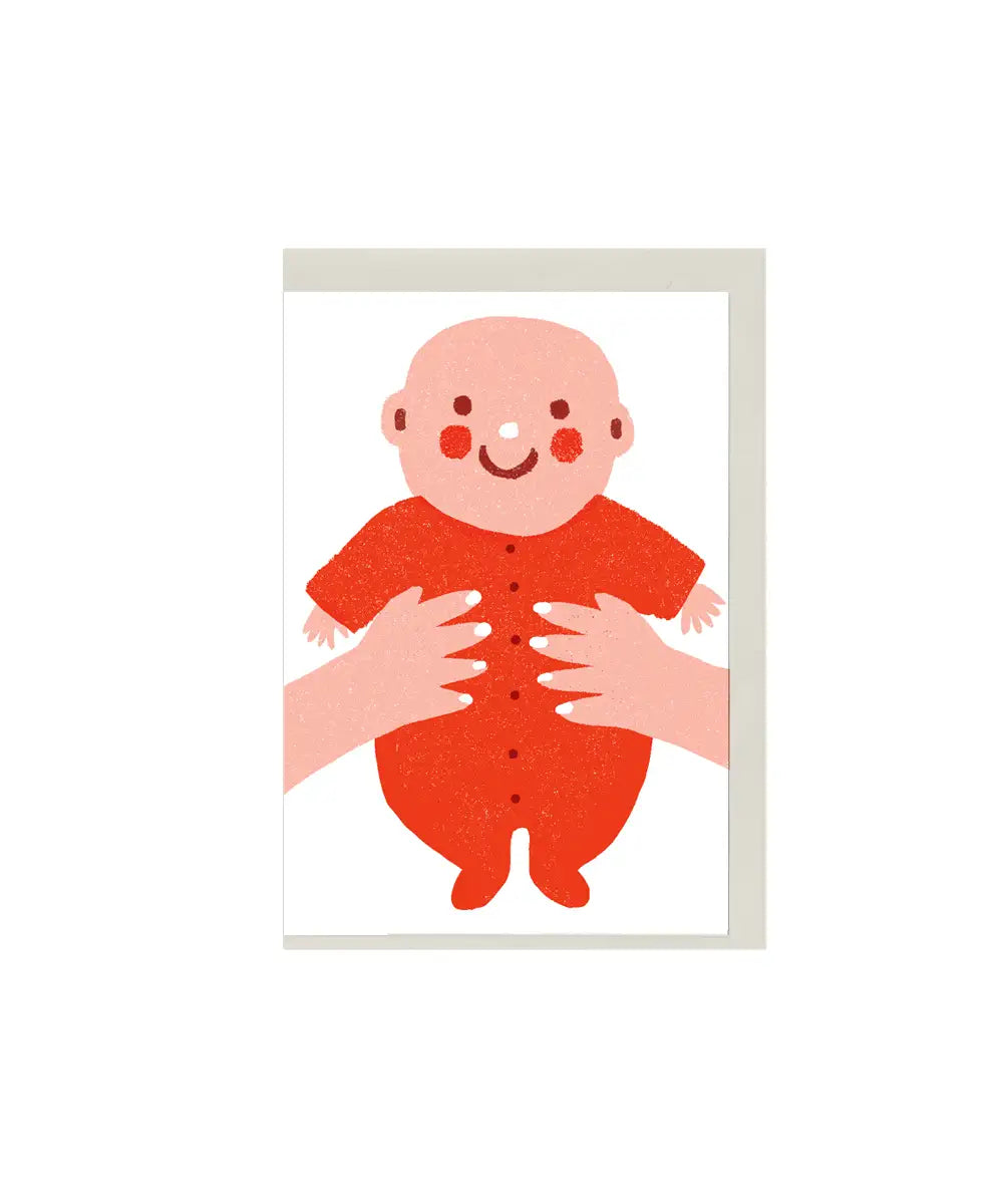 Illustrated new baby greeting card with smiling baby in red outfit, held by hands, mini A7 baby gift card by The Printed Peanut
