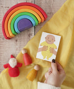 The Printed Peanut yellow new baby mini greetings card shown in an adult's hand surrounded by wooden toys in the Babipur playroom.