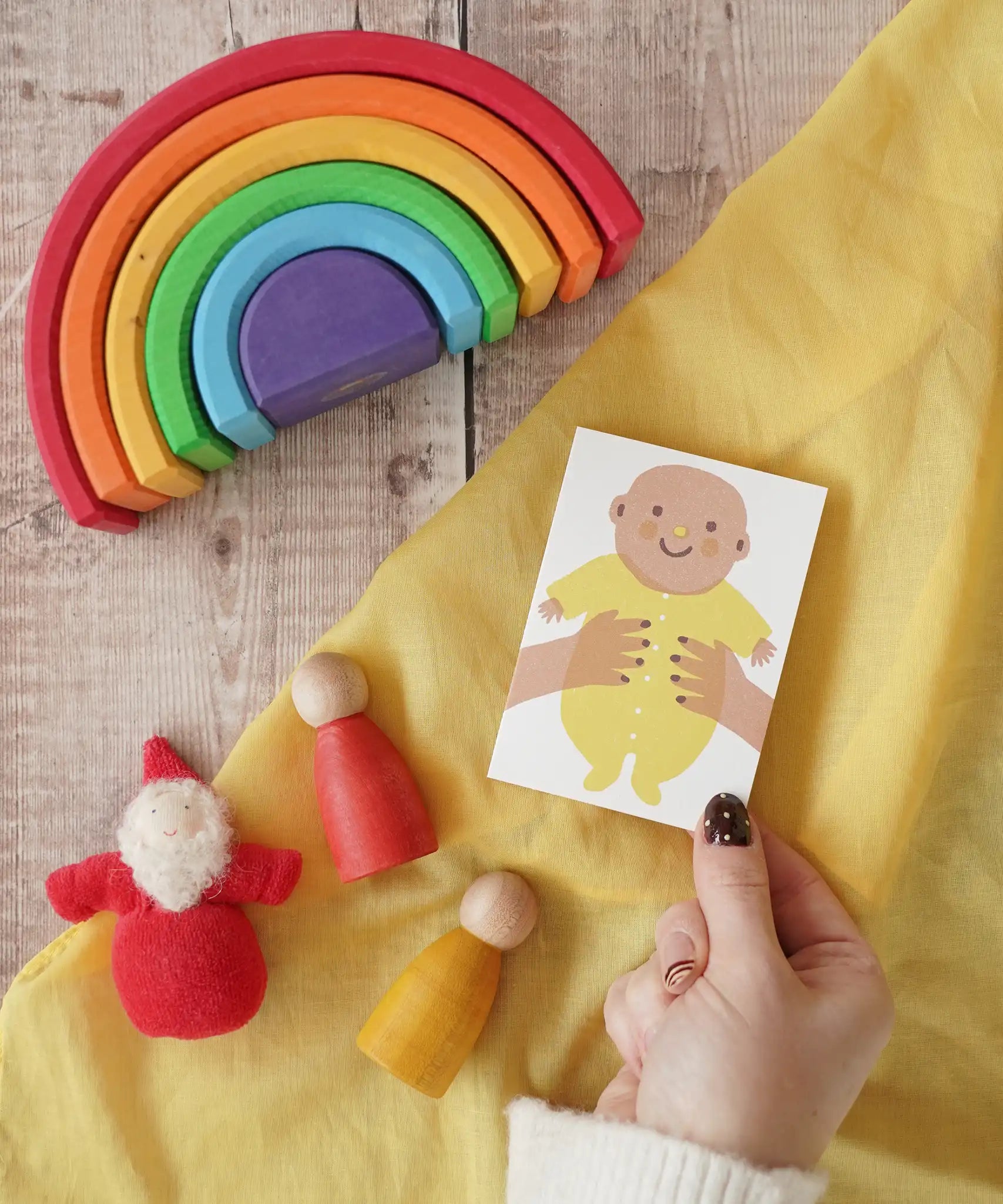 The Printed Peanut yellow new baby mini greetings card shown in an adult's hand surrounded by wooden toys in the Babipur playroom.