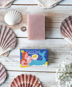 Printed Peanut rose geranium natural soap bar placed on a white wooden surface next to it's mermaid themed illustrated packaging box surrounded by seashells.