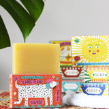 Printed peanut ylang ylang natural soap bar placed on top of the cardboard packaging box. Red box with dalmatian style dog illustration on the front. Other soaps from the collection can be seen in the background. 