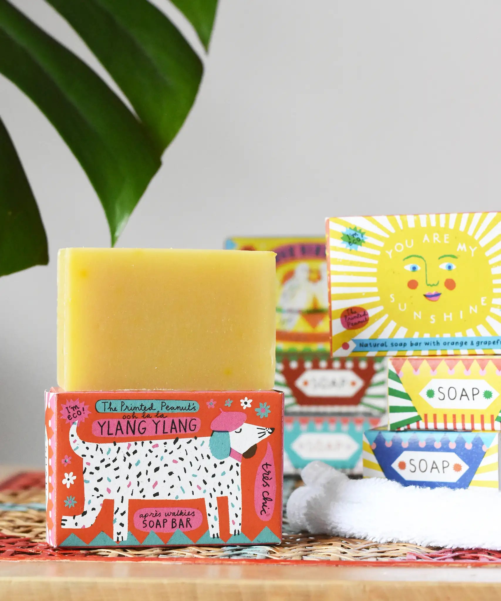 Printed peanut ylang ylang natural soap bar placed on top of the cardboard packaging box. Red box with dalmatian style dog illustration on the front. Other soaps from the collection can be seen in the background. 