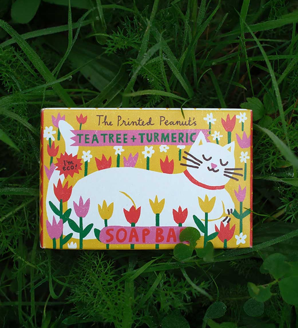 The Printed Peanut tea tree & turmeric natural handmade soap bar in a decorative cardboard packaging box. The box has been placed on grass.