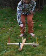A child kneeling, playing with the Wooden Story Ring Toss Game. One of the hoops can be see mid-air about to land on one of the coloured wooden posts.