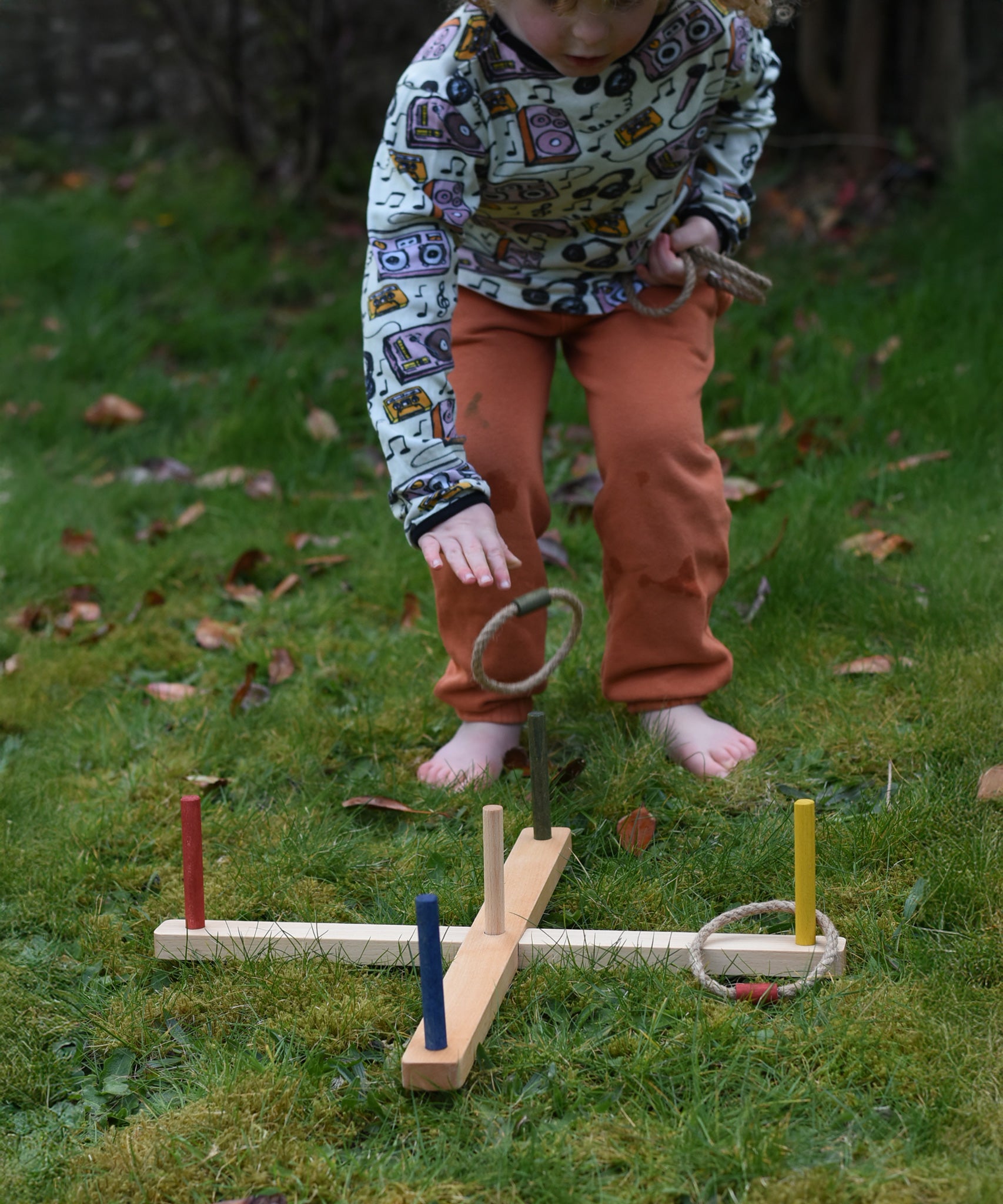 A child kneeling, playing with the Wooden Story Ring Toss Game. One of the hoops can be see mid-air about to land on one of the coloured wooden posts.