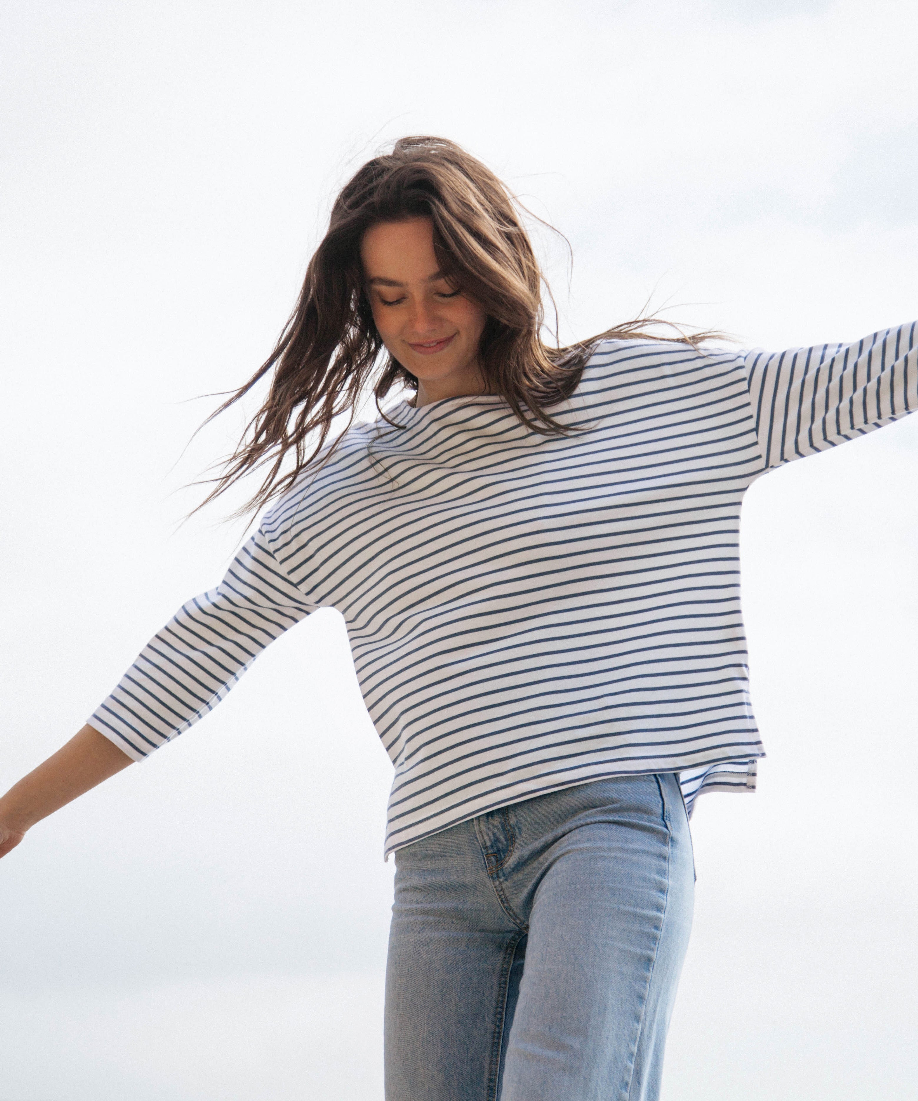 A woman standing outside wearing the Rapanui striped boat neck t-shirt in solent stripe design. A relaxed fit style organic cotton long sleeve top with a blue and white stripe design