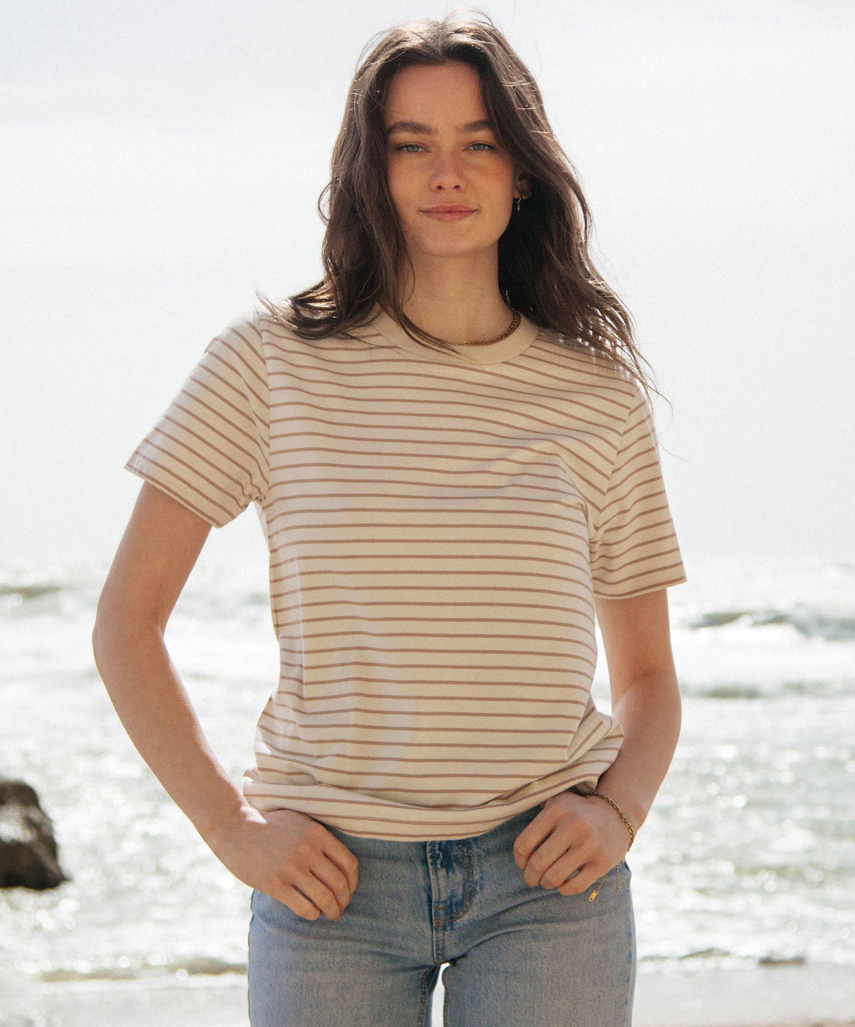 A woman standing outside wearing the Rapanui striped t-shirt in a taupe stripe design. A organic cotton short sleeve top in a relaxed fit style. 