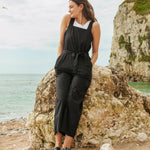 A woman sitting on a boulder on the beach wearing the black Rapanui jumpsuit. A organic cotton outfit with shoulder straps and a matching black belt tie