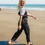 A woman walking on the beach wearing the black Rapanui organic cotton jumpsuit and striped top. 