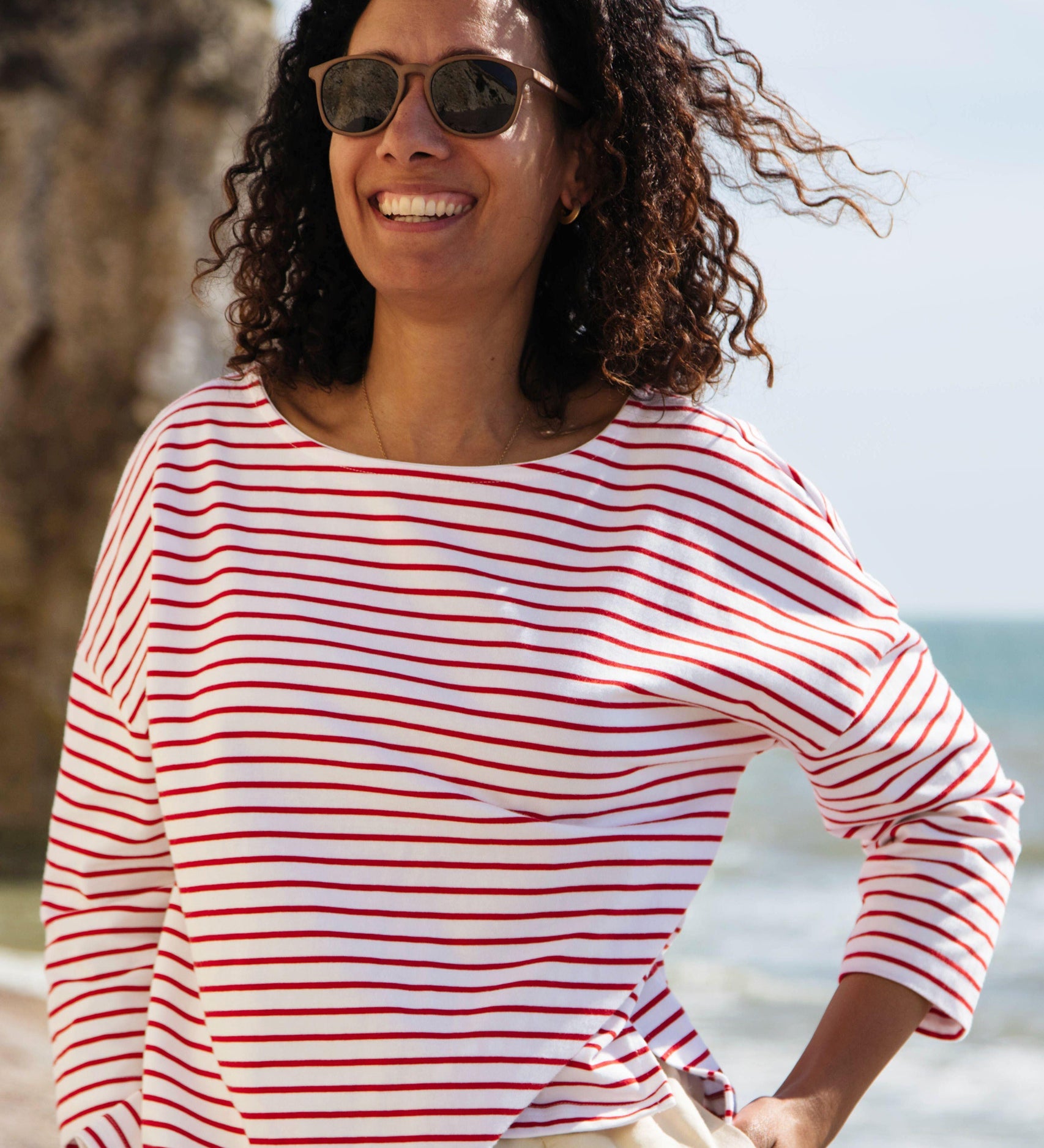 A woman standing outside wearing the Rapanui striped boat neck t-shirt in redwood stripe design. A organic cotton long sleeve top in a relaxed fit style with white and red stripes