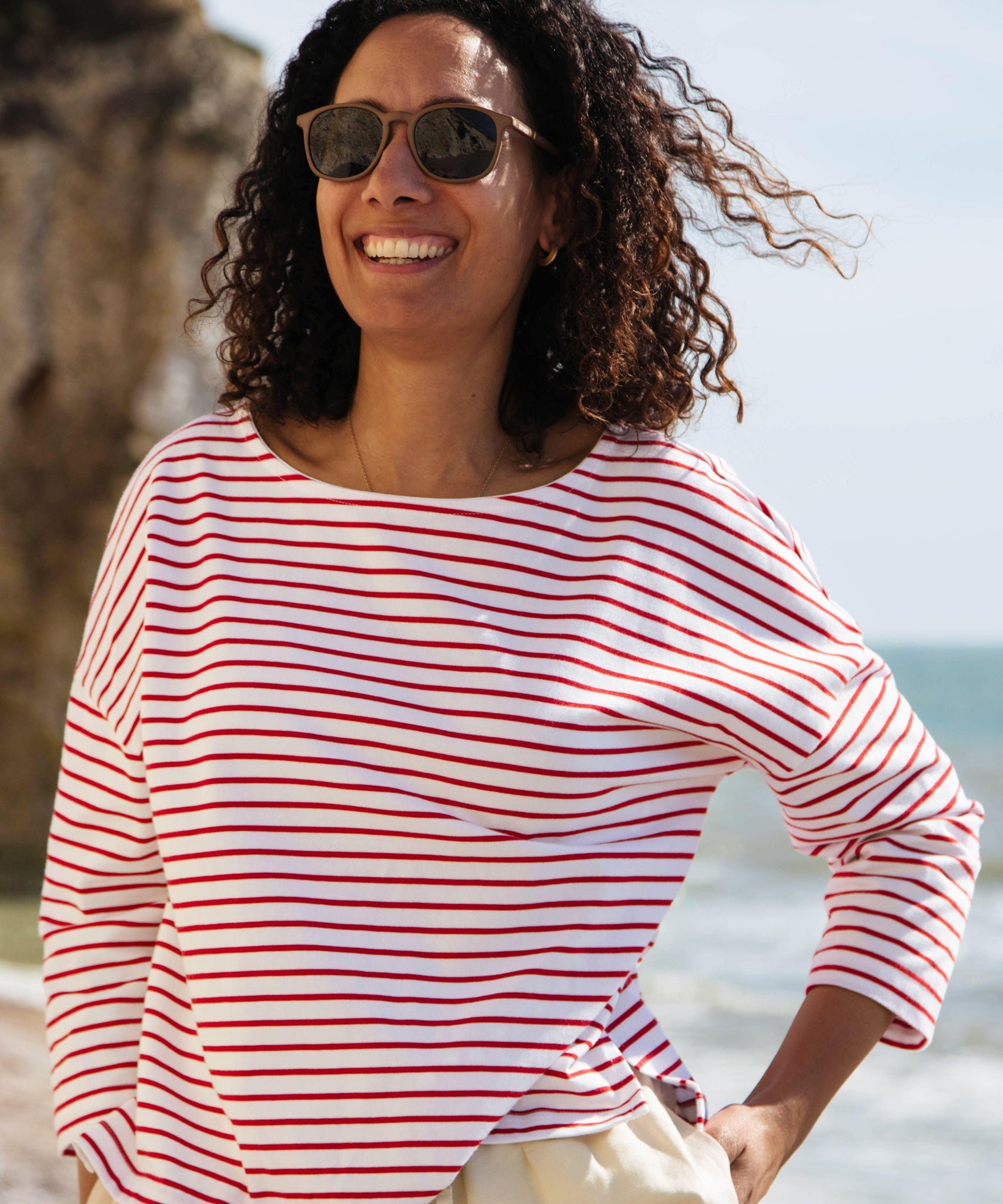 A woman standing outside wearing the Rapanui striped boat neck t-shirt in redwood stripe design. A organic cotton long sleeve top in a relaxed fit style with white and red stripes