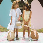 A child smiling and holding an Olli Ella Lamby Doll. In front of the child is an Olli Ella Rattan Lined Basket in Gumdrop