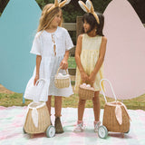 A child smiling and holding an Olli Ella Lamby Doll. In front of the child is an Olli Ella Rattan Lined Basket in Gumdrop