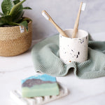 Rawr Shell Terrazzo Round Storage Pot in a bathroom setting. The pot holds two toothbrushes and sits next to a soap bar