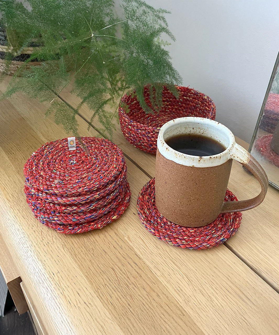 6 Respiin recycled multicolured coasters next to the matching holder on top of a wooden table with a mug on top of a coaster