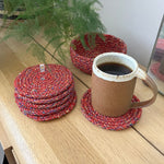 6 Respiin recycled multicolured coasters next to the matching holder on top of a wooden table with a mug on top of a coaster