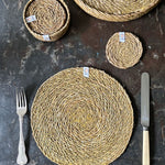 A top down view of the Respiin seagrass tablemat next to matching coasters on top of a stone surface next to a matching coasters 