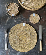 A top down view of the Respiin seagrass tablemat next to matching coasters on top of a stone surface next to a matching coasters 