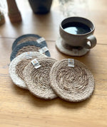 Set of 6 ReSpiin natural jute coasters on  a wooden table with a cup of coffee standing on one coaster