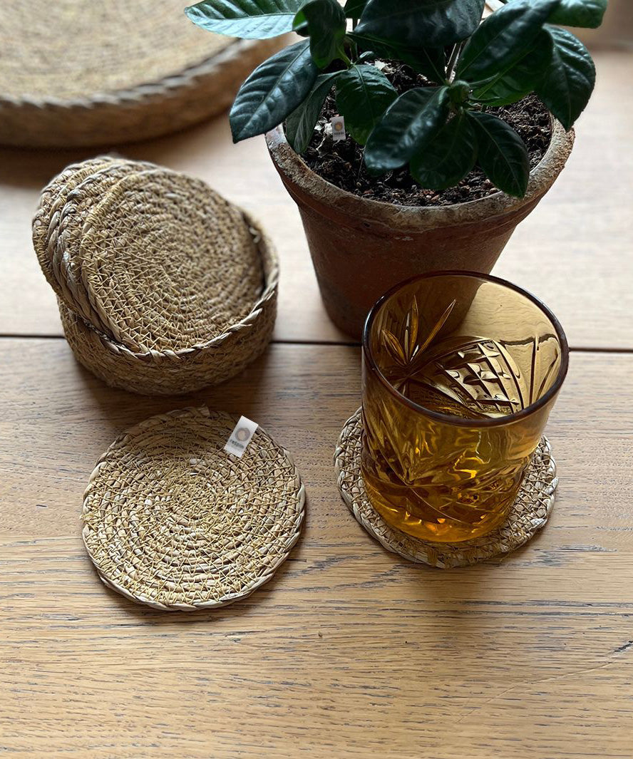 A close up of the 6 Respiin seagrass coasters in natural with a matching holder on top of a wooden table with a glass on top of a coaster
