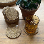 A close up of the 6 Respiin seagrass coasters in natural with a matching holder on top of a wooden table with a glass on top of a coaster