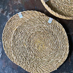 A top down view of the 6 Respiin seagrass tablemat in natural with a matching holder on top of a stone surface Features a brand patch stitched on the rim.